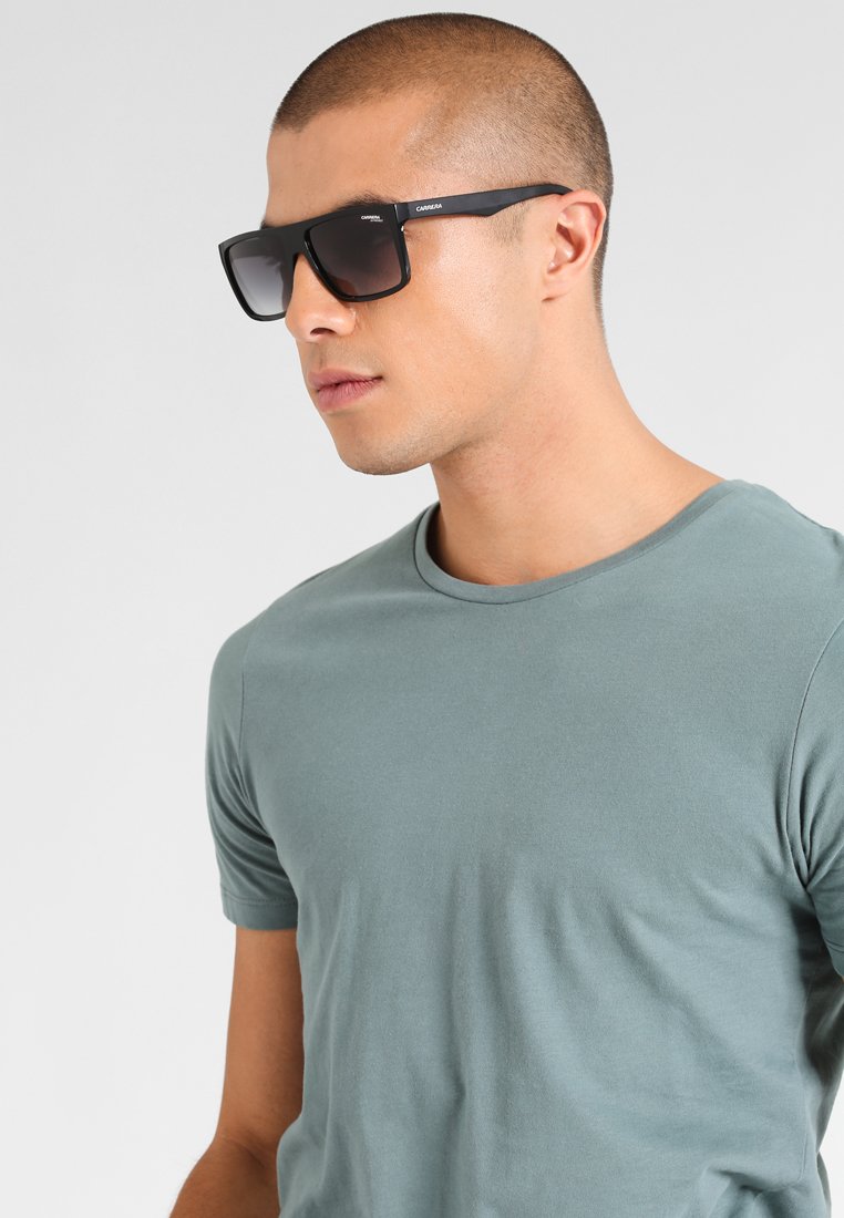 Black sunglasses with a rectangular frame, dark lenses, and subtle branding. The background is neutral gray. The model wears a light green shirt.