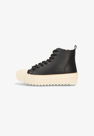 Zapatilla alta negra con suela zigzag gruesa beige, puntera beige, cordones negros y tirador sobre fondo blanco.