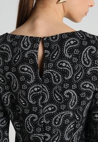 Tessuto nero con motivo paisley bianco, caratterizzato da un'apertura a chiave sul retro. Texture liscia ed elementi di design strutturati.