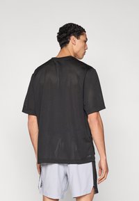 Μαύρο, κοντομάνικο δίχτυ μπλουζάκι με φαρδιά γραμμή, στρογγυλή λαιμόκοψη και μικρό λογότυπο κοντά στο τελείωμα. Το ύφασμα έχει διαπνεύσιμη υφή.