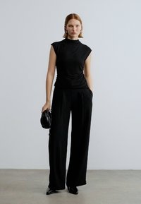 Top preto sem mangas com gola alta, combinado com calças pretas de perna larga. Modelo segura uma pequena bolsa preta. Textura suave, design ajustado.