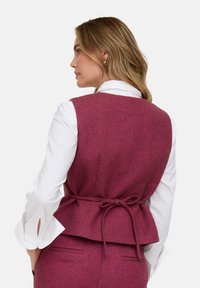 Weinrotes Fischgrätenmuster-Weste mit figurbetonter Silhouette, verstellbarer Bindung am Rücken, getragen über einer weißen Bluse mit Manschetten-Schlitzen. Keine sichtbaren Verschlüsse.