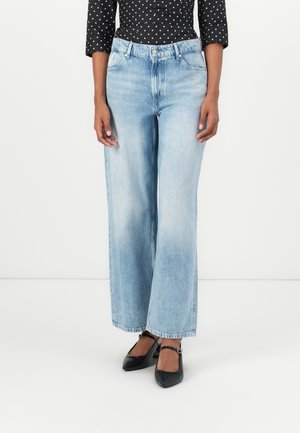 Kobieta ubrana w jasnoniebieskie jeansy o szerokich nogawkach, czarne spiczaste buty Mary Jane oraz czarną bluzkę w białe kropki, stojąca na białym tle.