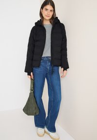 Sort pufferjakke med hætte, grå langærmet top, blå flared jeans og en grøn quiltet taske. Hvide sneakers med beige såler.
