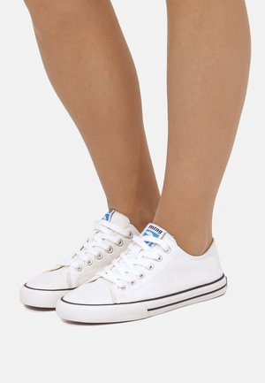 Gambe inferiori di una persona che indossano sneakers basse in tela bianca con lacci bianchi e suole in gomma a strisce nere su uno sfondo semplice.