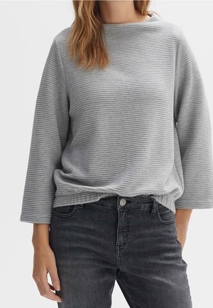 Femme portant un haut à manches longues côtelé gris clair avec un jean noir sur un fond blanc uni.