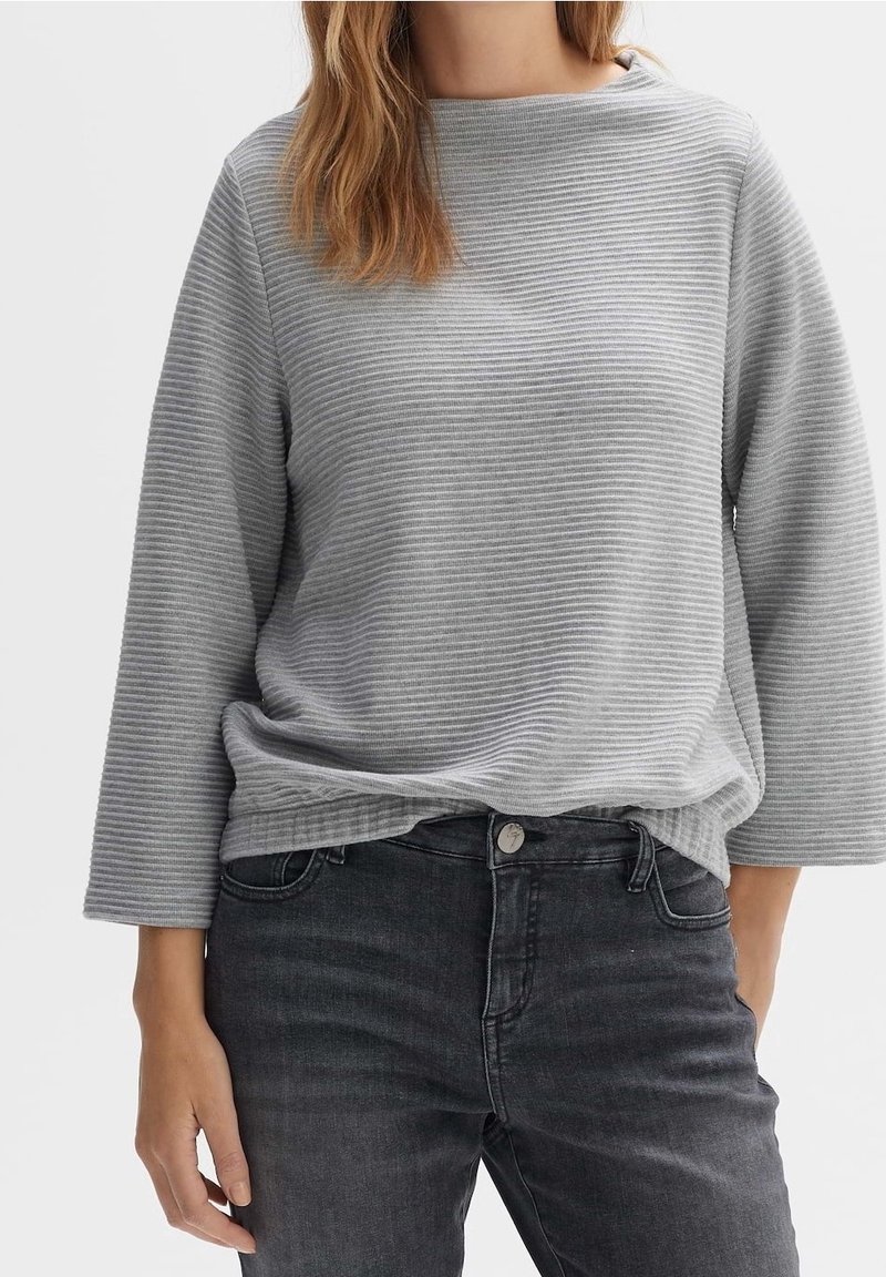 Femme portant un haut à manches longues côtelé gris clair avec un jean noir sur un fond blanc uni.