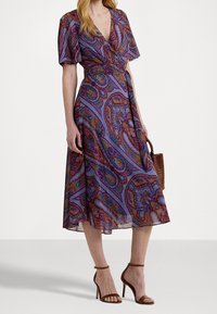 Robe froncée violette à manches courtes, motif cachemire dans des couleurs vives, descend jusqu'au mollet, complétée par des talons à lanières marron et un sac en osier.