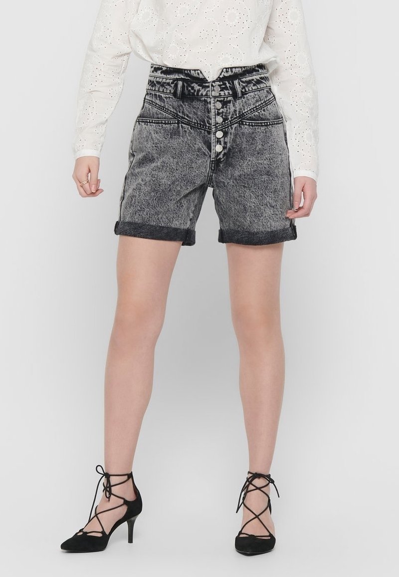 Personne portant un chemisier blanc à œillets, un short en jean noir taille haute effet délavé avec boutons et des talons noirs à lacets, debout.