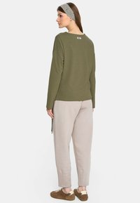 Oliwkowa bluzka z długim rękawem i okrągłym dekoltem, zestawiona z jasnobeżowymi spodniami o tapered fit. Stylizacja uzupełniona jest teksturowaną opaską na głowę i brązowymi butami.