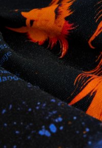 Bavlněná tkanina s designem oranžového lva na černém pozadí, s modrým textem a barevnými tečkami různých velikostí a odstínů.