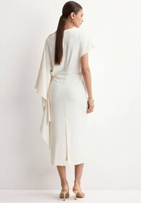 Woman with long hair in a white midi dress featuring an asymmetrical draped sleeve and back slit, wearing beige heeled sandals and gold jewelry.