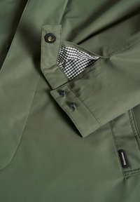 Chaqueta impermeable verde oliva con botones a presión negros y un forro de cuadros. Presenta costuras diagonales y una etiqueta en el bolsillo.
