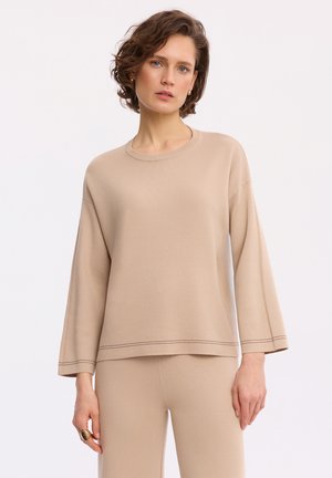 Kobieta z krótkimi kręconymi włosami, ubrana w beżową bluzkę z długim rękawem i pasujące spodnie, stojąca na białym tle.