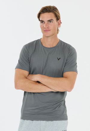 Jeune homme les bras croisés, portant un t-shirt de sport gris à manches courtes et un short gris clair sur un fond blanc uni.