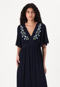 Navy maxi dress with a deep V-neck, featuring blue floral embroidery on the chest and short flutter sleeves; soft fabric and loose fit.