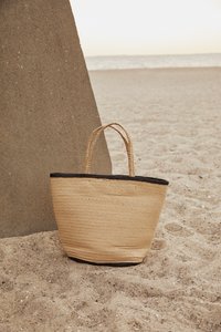 Sac cabas en paille tissée avec bordure noire reposant contre une grosse pierre sur une plage de sable près d’une mer calme.