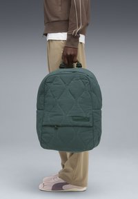 Zaino verde cucito a quadri con cuciture curve, manici imbottiti e tasca frontale. Indossato da una persona con pantaloni beige e sneakers rosa.