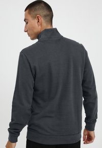 Temno siva kapuca s zadrgo z visokim ovratnikom, rebrastimi manžetami in robom. Mehak material z minimalnim logotipom vidnim na strani.