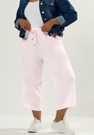 Kvinde iført lyserøde, vidde, cropped bukser med snøre, hvide sneakers, hvid top og blå denimjakke mod en neutral baggrund.