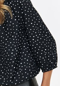 Czarna bluzka w białe kropki z bufiastymi rękawami i marszczonymi mankietami. Miękka tkanina w luźnym fasonie.