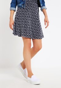 Kvinde iført et sort og hvidt mønstret nederdel, denimjakke og hvide slip-on sko, stående med det ene ben krydset over det andet.