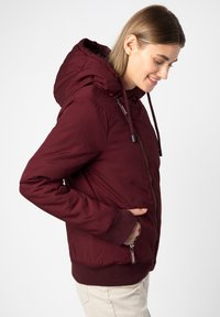 Maroon padded jacket with a hood, elastic cuffs, side pockets, and a front zipper. Smooth fabric with a slight sheen and structured fit.