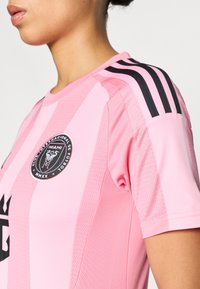 Világos rózsaszín focimez fekete csíkokkal a vállakon, kör alakú fekete emblémával, rajta a „MIAMI” felirattal és mintás textúrával.