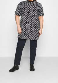 Svart och vit t-shirtklänning med textmönster "CALVIN KLEIN JEANS". Korta ärmar, avslappnad passform, ihopparad med marinblå byxor och svarta ankelskor.