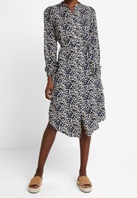 Klänning med leopardmönster i kräm, marinblå och beige. Långa ärmar, krage, midjeband, asymmetrisk nederkant, lättviktsmaterial.