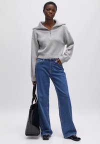 Lichtgrijze rits trui met een brede kraag, gecombineerd met hoge taille blauwe jeans en een zwarte tote bag, tegen een neutrale achtergrond.