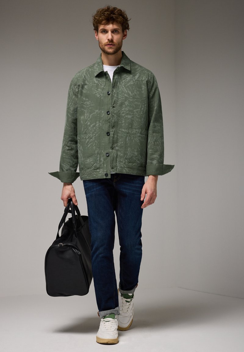 Zelená vzorovaná bunda s černými knoflíky, nošená přes bílý tričko, v kombinaci s tmavými džínami a bílými teniskami, nese černou tašku.