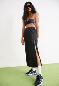 Fekete sportos szett, amely egy cropped bandeau topból és egy hosszú szoknyából áll, oldalsó cipzárral. Fekete sportcipővel és fehér zoknikkal viselték.