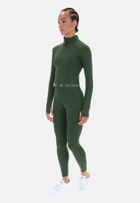 Olívazöld atlétikai szett hosszú ujjakkal és magas nyakkal; testhezálló kialakítás, fehér "TED BAKER" feliratos szalaggal a deréknál; fehér sportcipővel viselve.