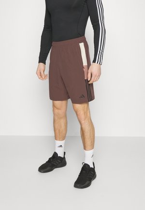 Mann trägt braune Shorts mit Seitenstreifen, ein schwarzes Langarmshirt mit weißen Arstreifen, weiße Adidas-Socken und schwarze Adidas-Sneaker.