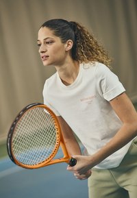 White cotton t-shirt with orange "BJÖRN BORG" branding, paired with olive green pants. Holding an orange tennis racket with a textured grip.