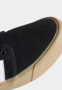 Černá semišová teniska s texturovaným povrchem, vybavená podrážkou z gumy barvy žvýkačky a bílým proužkem podél strany. Zaoblená špička.