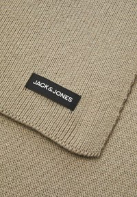 Bežani pleteni šal s teksturiranim uzorkom i crnom etiketom s natpisom "JACK & JONES" u bijelom slovu.