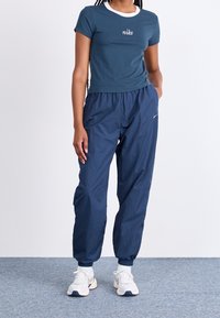 Mladý člověk oblečený v tmavě modrém tričku Nike, odpovídajících teplákových kalhotách a bílých teniskách, stojící na šedém koberci.