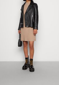 Veste de motard en cuir noir avec des zips, robe en tricot beige et bottes noires à lacets épaisses avec des lacets jaunes, accompagnées d'un petit sac à main noir.