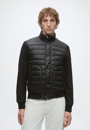 Jeune homme aux cheveux bouclés portant une veste matelassée noire avec des poches zippées et un pantalon blanc, debout devant un fond clair uni.
