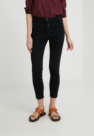 Frau trägt schwarze, verkürzte Skinny-Jeans, bordeauxfarbenes Hemd mit hochgekrempelten Ärmeln und braune sowie goldene Sandalen mit offenen Zehen, steht auf weißem Boden.