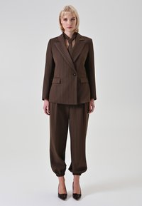 Completo marrone a righe sottili con blazer doppiopetto e pantaloni stretti. Il blazer presenta un bottone singolo; i pantaloni hanno polsini elasticizzati.