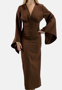 Vestito marrone a maniche lunghe, con scollo a V profondo, silhouette aderente e maniche svasate realizzate in tessuto liscio. Lunghezza fino a metà polpaccio.