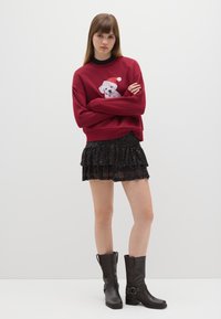 Rode sweatshirt met een hondenafbeelding die een Santa-hat draagt, gecombineerd met een zwarte glitter ruffle minirok en bruine enkellaarsjes.