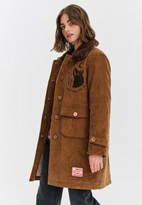 Brązowy sztruksowy płaszcz z futrzanym kołnierzem, z haftowanym wzorem, z zapięciem na guziki oraz naszywką w dolnym rogu.