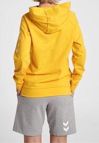 Sudadera amarilla con capucha y mangas largas, que cuenta con un bolsillo frontal y detalles de logotipo en blanco. Combinada con pantalones cortos grises que presentan un patrón de chevron.
