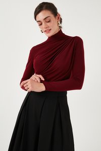 Žena s tmavými vlasmi, ktorá nosí zlaté kruhové náušnice, bordový dlhý nariasený top a čiernu plisovanú sukňu, pózujúc so skríženými rukami.