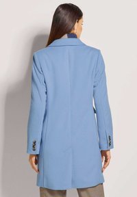 Helleblaue Wolljacke mit maßgeschneidertem Design, langen Ärmeln, Rückenschlitz und seitlichen Taschen, mit Knopfdetails an den Bündchen.