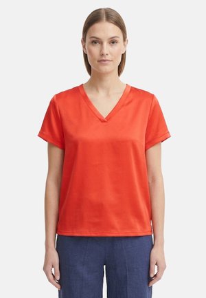 Donna con capelli lisci che indossa una maglietta arancione a maniche corte con scollo a V e pantaloni blu, in piedi davanti a uno sfondo uniforme.